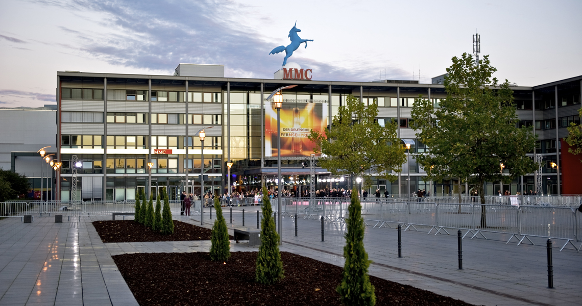 Außenansicht des Haupteingangs des MMC Coloneum in Köln am Abend. Zu sehen sind der moderne Gebäudekomplex, der gepflasterte Vorplatz mit jungen Bäumen, die markante blaue Pegasus-Skulptur und das MMC-Logo auf dem Dach.